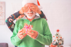 Senior couple in warm clothing and santa hat, woman covering eyes of old husband with a surprise gift for him. Loving old romantic heterosexual couple celebrating christmas festival together